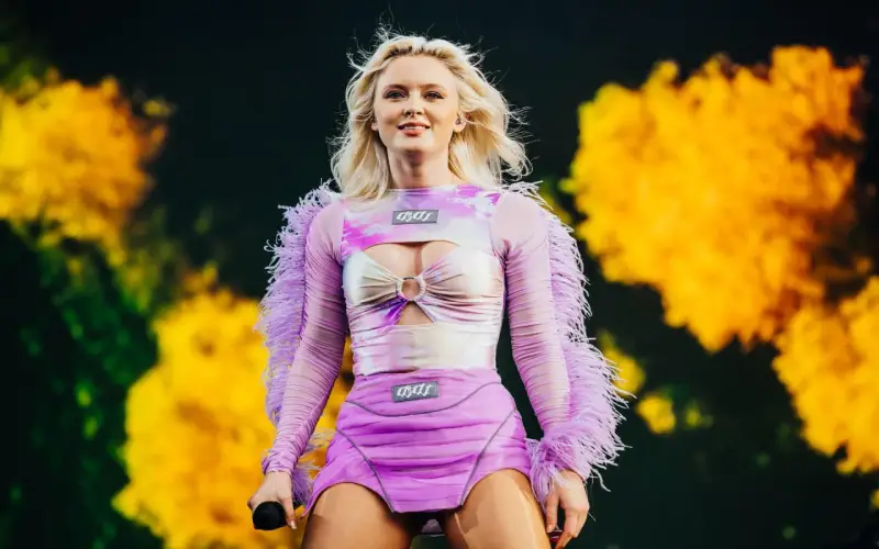 Zara Larsson konsert. Hon står på scenen i en lila och rosa outfit med fjädrar på ärmarna.