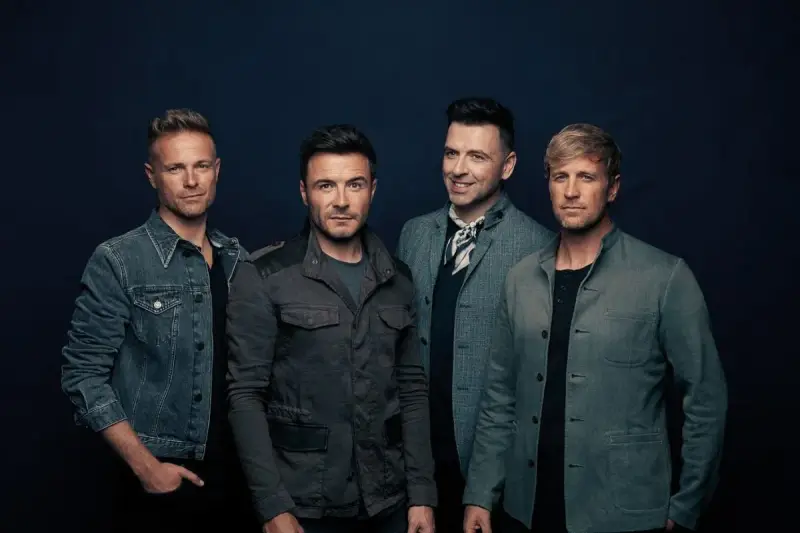 Medlemmar av Westlife poserar för en studiofotografering. De bär moderna jackor och ser direkt in i kameran.